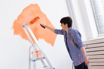 Pretty smiling middle-aged woman painting interior wall of home with paint roller. Redecoration, renovation, apartment repair and refreshment concept.