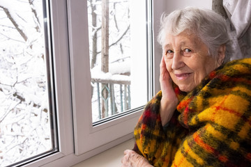 Old lonely woman sitting near the window in his house.