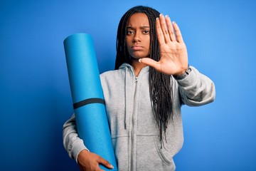 Young african american fitness woman holding yoga mat over isolated blue background with open hand...