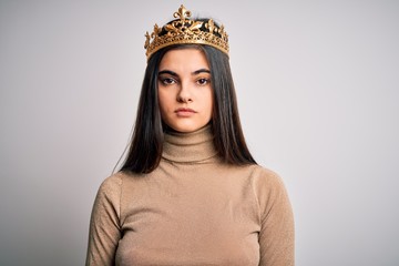 Young beautiful brunette woman wearing golden queen crown over isolated white background with serious expression on face. Simple and natural looking at the camera.