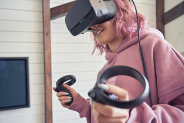Hipster gamer teen girl pink hair wear vr headset goggles hold handheld controllers play video game...