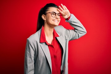 Young beautiful brunette businesswoman wearing jacket and glasses over red background very happy and smiling looking far away with hand over head. Searching concept.