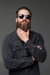 Young handsome man in sunglasses touch his long beard isolated on gray background