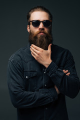 Young handsome man in sunglasses touch his long beard isolated on gray background