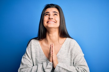 Obraz premium Young beautiful brunette woman wearing casual sweater standing over blue background begging and praying with hands together with hope expression on face very emotional and worried. Begging.