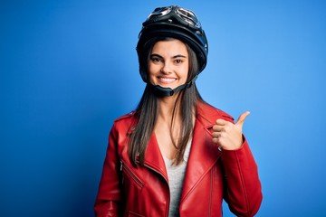 Young beautiful brunette motorcycliste woman wearing motorcycle helmet and jacket smiling with happy face looking and pointing to the side with thumb up.