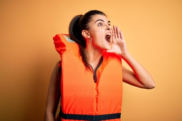 Young beautiful brunette woman wearing orange safe lifejacket over yellow background shouting and...