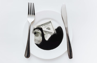 A hundred-dollar bill lies in a white plate near the fork, knife and is filled with black oil. The...