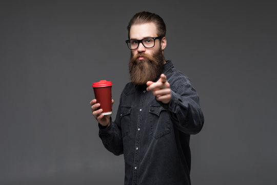 Handsome bearded man with stylish hair beard and mustache on serious face pointed on camera holding cup or mug drinking tea or coffee on grey background