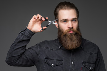 Young bearded man holding barber scissors and razor near his face standing isolated on gray background