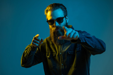 Young hipster bearded man pointed on camera wearing sunglasses isolated on light background