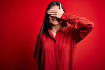 Young beautiful woman with blue eyes wearing casual shirt standing over red background smiling and laughing with hand on face covering eyes for surprise. Blind concept.