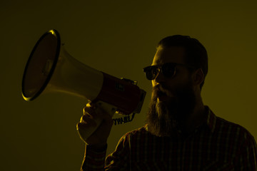 Young hipster man shouting by megaphone isolated on yellow background