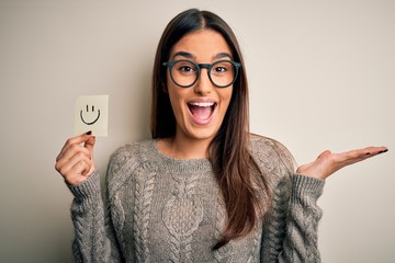 Young beautiful brunette woman wearing glasses holding paper with smile emoji very happy and excited, winner expression celebrating victory screaming with big smile and raised hands
