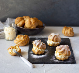 Cream puffs or profiteroles with whipped cream dusted with sugar powder on slate board, filling split puff with cream using knife