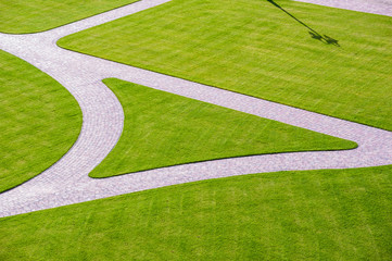 Cobbled park alleys surrounded by green grass on a sunny, bright spring day. Lamp shade visible on grass. Park seen from above.