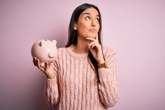 Young Beautiful Brunette Woman Holding Piggy Bank Saving Money For Retirement Serious Face Thinking About Question, Very Confused Idea