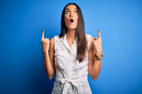 Young beautiful brunette woman wearing casual dress over isolated blue background amazed and surprised looking up and pointing with fingers and raised arms.