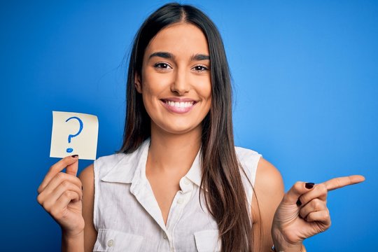Young beautiful brunette woman holding paper with question mark symbol message very happy pointing with hand and finger to the side