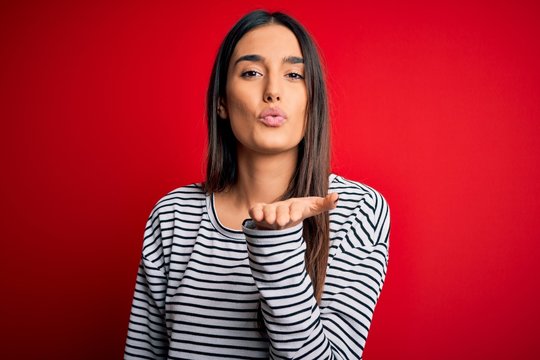 Young beautiful brunette woman wearing casual striped t-shirt over red background looking at the camera blowing a kiss with hand on air being lovely and sexy. Love expression.
