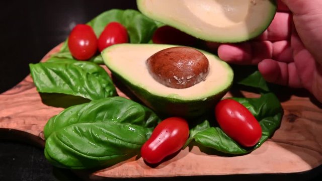 A wooden cutting board on a black background with an avocado in the center. All around, intense green basil leaves and bright red cherry tomatoes. One hand raises the half doing revealing the inside