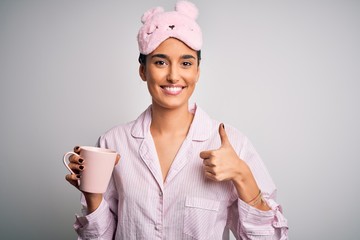 Young beautiful brunette woman wearing pajama and sleep mask drinking cup of coffee happy with big smile doing ok sign, thumb up with fingers, excellent sign