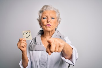 Senior beautiful grey-haired woman holding reminder paper heart over white background pointing with...