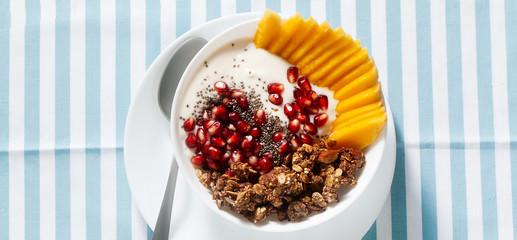banner of breakfast bowl of granola, yogurt, fresh sweet mango, chia seeds, pomegranate seeds and maple syrup on a blue striped napkin