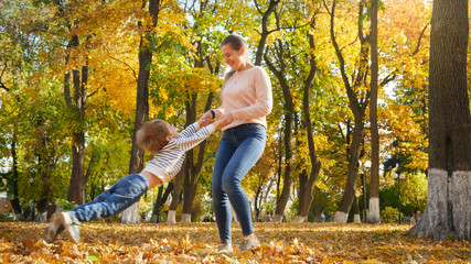 Young msiling woman holding her son by hands and spinning him at autumn park