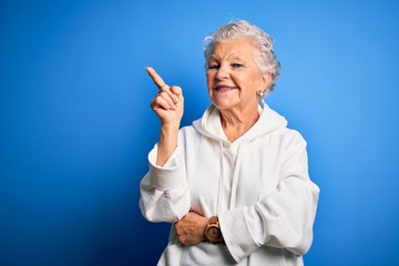 Senior beautiful sporty woman wearing white sweatshirt over isolated blue background with a big smile on face, pointing with hand and finger to the side looking at the camera.