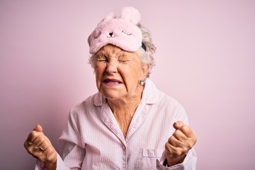 Senior beautiful woman wearing sleep mask and pajama over isolated pink background very happy and excited doing winner gesture with arms raised, smiling and screaming for success. Celebration concept.