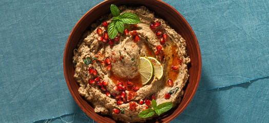banner of homemade spread of baked eggplant baba ganoush in a bowl with pomegranate seeds, lime, olive oil and lime slices on a blue linen tablecloth
