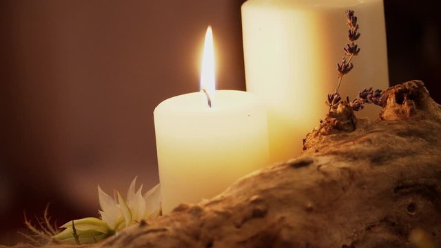 Flame of a white wax candle burns near a wooden bitch with flowers and lavender