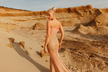 Rear view of a sensual young blonde woman in the desert in maxi gold dress posing in desert, during at sunset background.