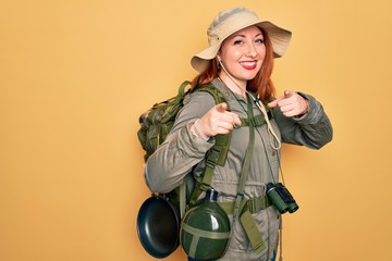 Young redhead backpacker woman hiking wearing backpack and hat over yellow background pointing...