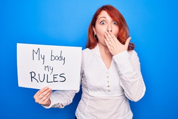 Young redhead woman asking for women rights holding paper with my body my rules message covering mouth with hand, shocked and afraid for mistake. Surprised expression