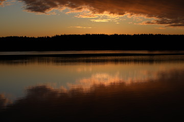 Mirror image of pink light sky with bright burning crimson clouds in the lake at dawn.The illuminated black forest on the horizon is copied in the calm water.A glossy picture in the twilight.Russia