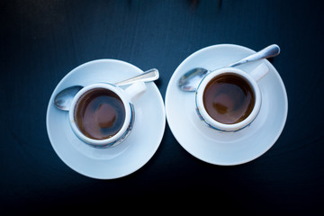 Two cups of coffee with round white plates and tea spoons on black table. View from above. Morning coffee in bar for a couple. Coffee espresso geometrical concept. Coffee-mania, black and white. 