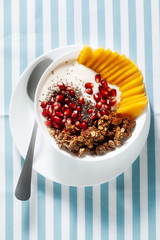 breakfast bowl of granola, yogurt, fresh sweet mango, chia seeds, pomegranate seeds and maple syrup on a blue striped napkin