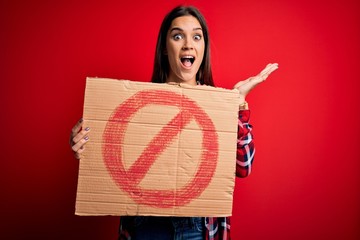 Young beautiful brunette woman holding banner with prohibited signal over red background very happy...