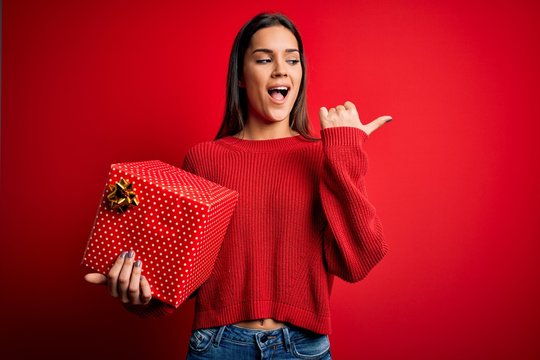 Young beautiful brunette woman holding birthday gift over isolated red background pointing and showing with thumb up to the side with happy face smiling