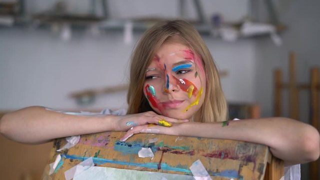 Portrait of a young attractive artist with paint on her face, leaning on an easel.