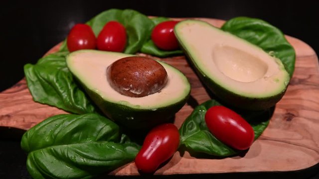 Static image of a wooden cutting board on a black background with an avocado cut in half in the center. All around, intense green basil leaves and bright red cherry tomatoes