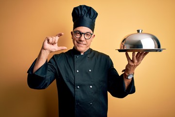 Middle age handsome grey-haired waiter man wearing cooker uniform and hat holding tray smiling and confident gesturing with hand doing small size sign with fingers looking and the camera. Measure