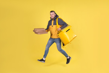 Fototapeta na wymiar Blond unshaven teen food delivery boy and courier in yellow clothes with thermo bag on his back holds an order for customer and runs to deliver order on time in 15 minutes. Food delivery in city