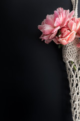 Crochet bag containing a bouquet of light pink peonies in the right part of a great black background.