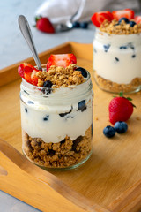 Healthy breakfast on wooden platter with containers containing natural yogurt with granola and berries. Strawberries, granola, blueberries, teaspoon.