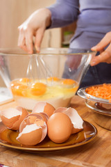 Skorupki kurzych jaj leżą na spodku na pierwszym planie. Na drugim planie kobieta roztrzepuje jajka kurze leżące w szklanej misce. Przygotowywanie ciasta. - obrazy, fototapety, plakaty