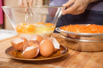 Skorupki kurzych jaj leżą na spodku na pierwszym planie. Na drugim planie kobieta roztrzepuje jajka kurze leżące w szklanej misce. Przygotowywanie ciasta.