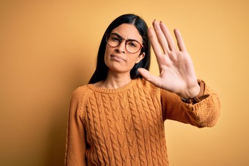 Young beautiful hispanic woman wearing glasses over yellow isolated background doing stop sing with...